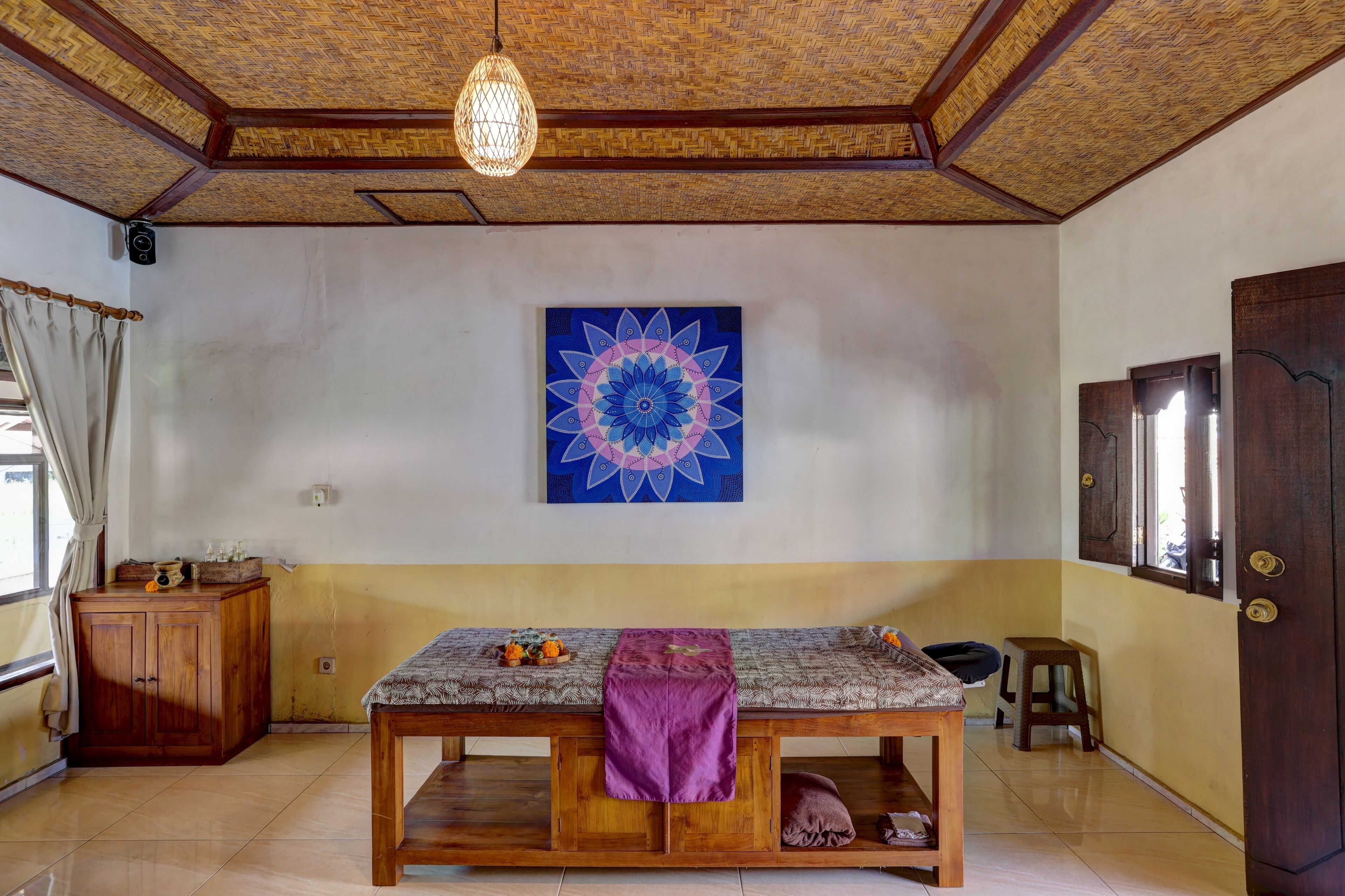Une pièce comprenant un banc en bois, un tissu violet, un mur orné d’un mandala bleu, une armoire en bois et une porte avec des fenêtres.