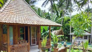 A wooden house with a tiled roof, a porch with chairs, and a pool area surrounded by tropical vegetation. - Junjungan Suite Villa (Ubud)