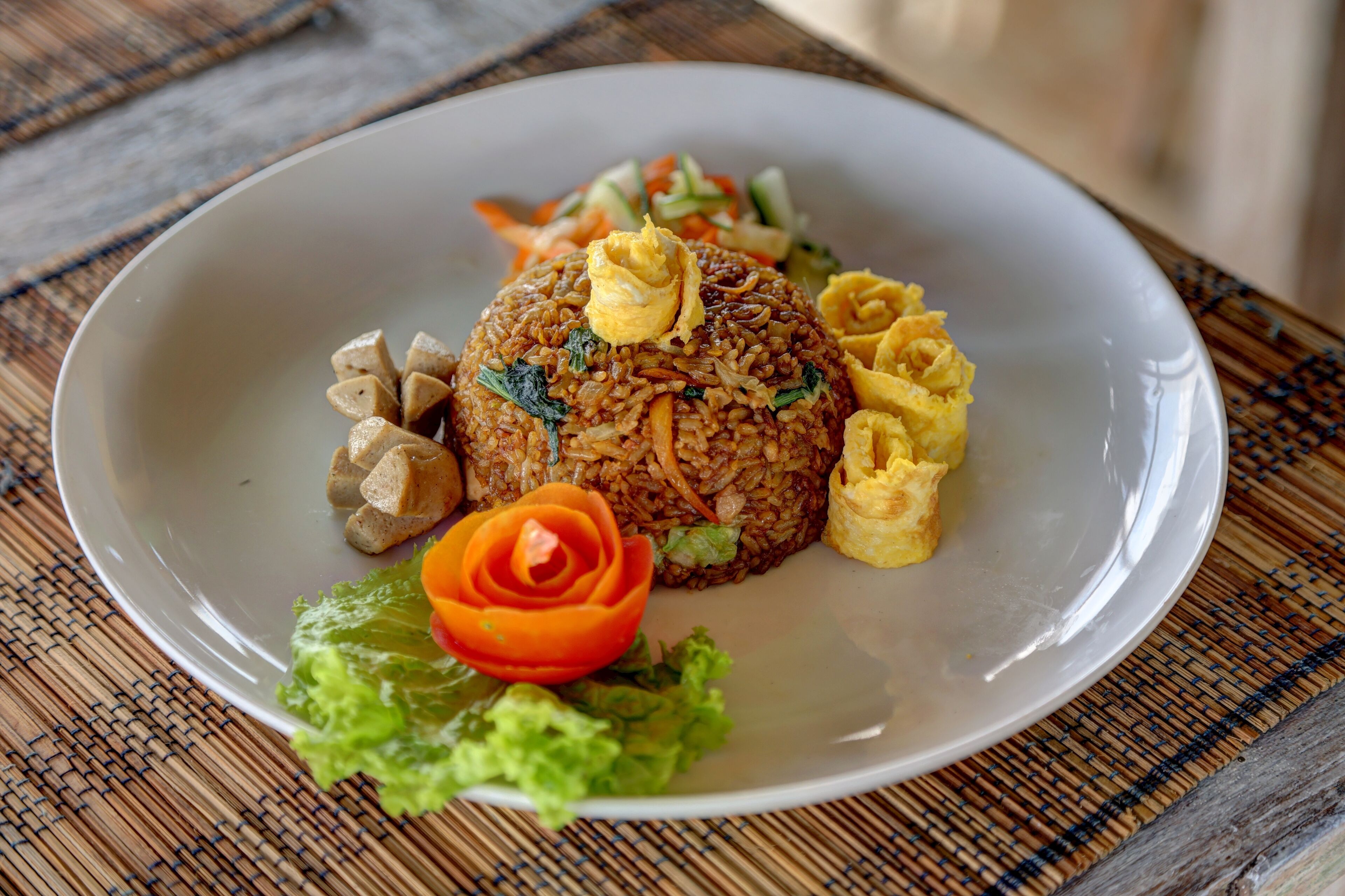 A plate of fried rice with vegetables and egg on a white plate, placed on a bamboo mat.