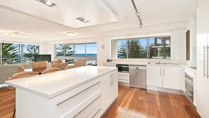 A modern kitchen with white cabinetry, a large island, and a view of the ocean. - Solnamara Beachfront Apartments (Burleigh Heads)