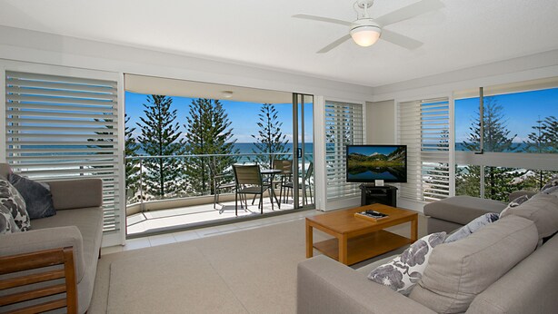 A modern living room with a television, sofa, and coffee table. There is a balcony with a view of trees and the ocean. - Solnamara Beachfront Apartments (Burleigh Heads)
