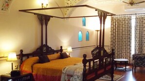 Una cama con dosel, suelo de madera, alfombra estampada y una silla en una habitación con papel tapiz floral y ventilador de techo.