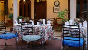 Un comedor con mesas dispuestas para cuatro, rodeado de columnas y arcos, con manteles florales y sillas de metal.