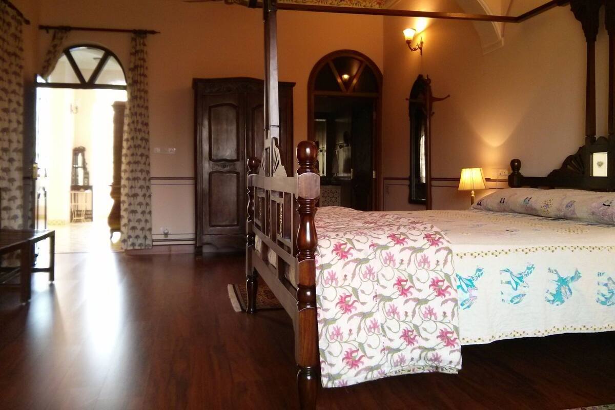 A bedroom with a four-poster bed, a wooden floor, a chandelier, and a view of the hallway.