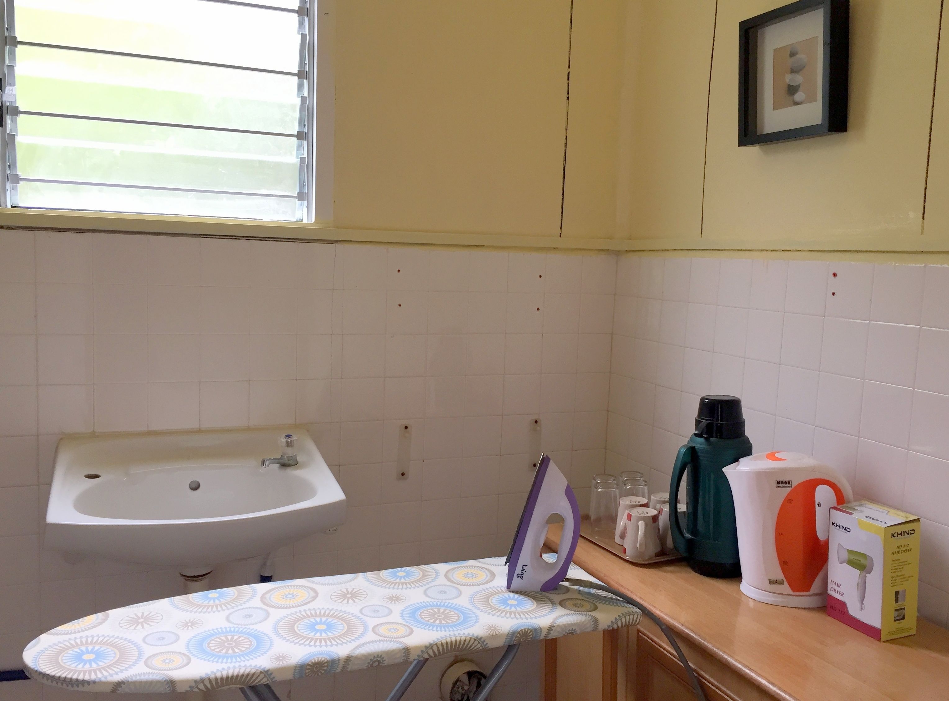 Een badkamer met een wastafel, een strijkplank, een haardroger en een ingelijste foto aan de muur.