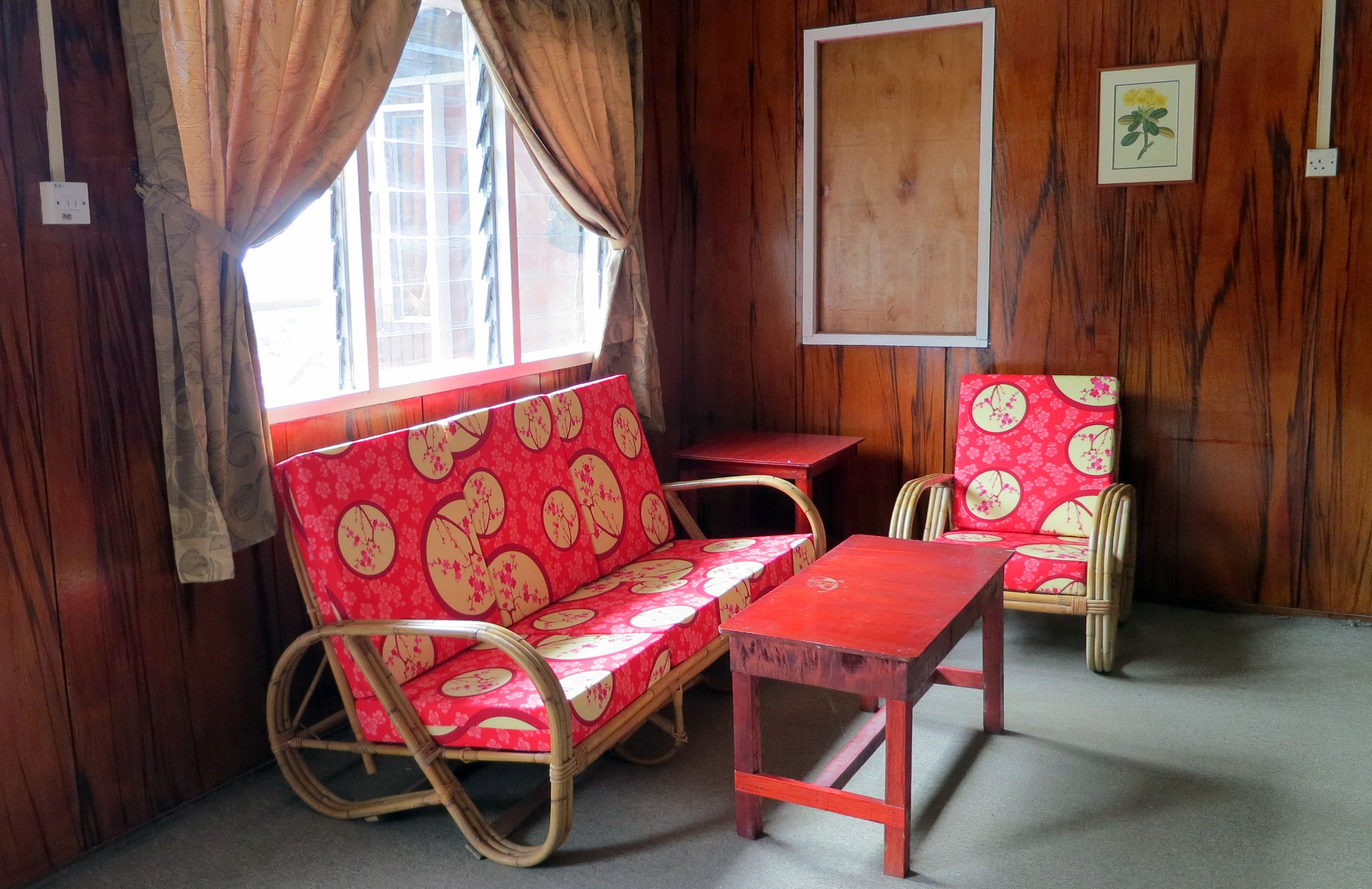 A room with wooden walls, a window with curtains, a red sofa with floral patterns, a matching armchair, and a small table.