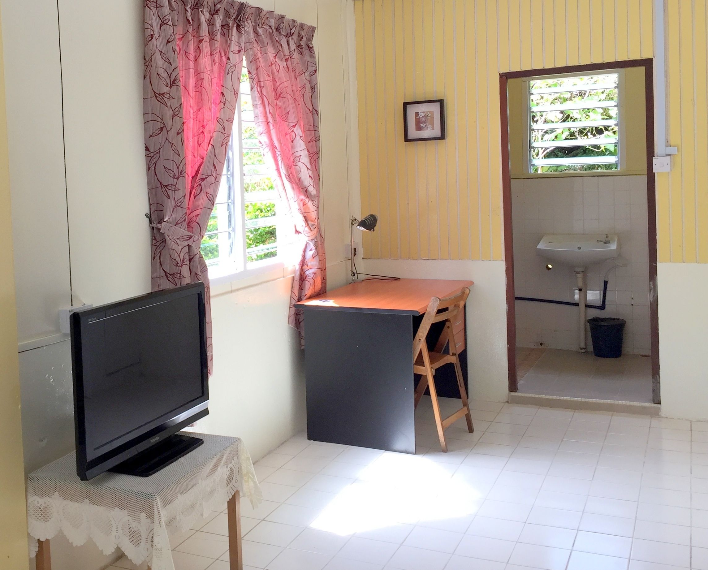 Quarto com televisão sobre uma mesa, secretária, cadeira, janela com cortinas, pia e porta que dá para uma varanda.
