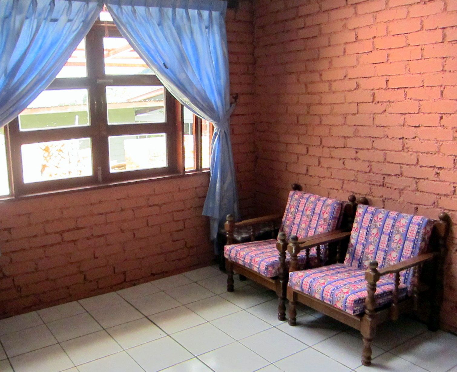 A room with a brick wall, two wooden chairs with patterned cushions, a window with blue curtains, and a tiled floor.