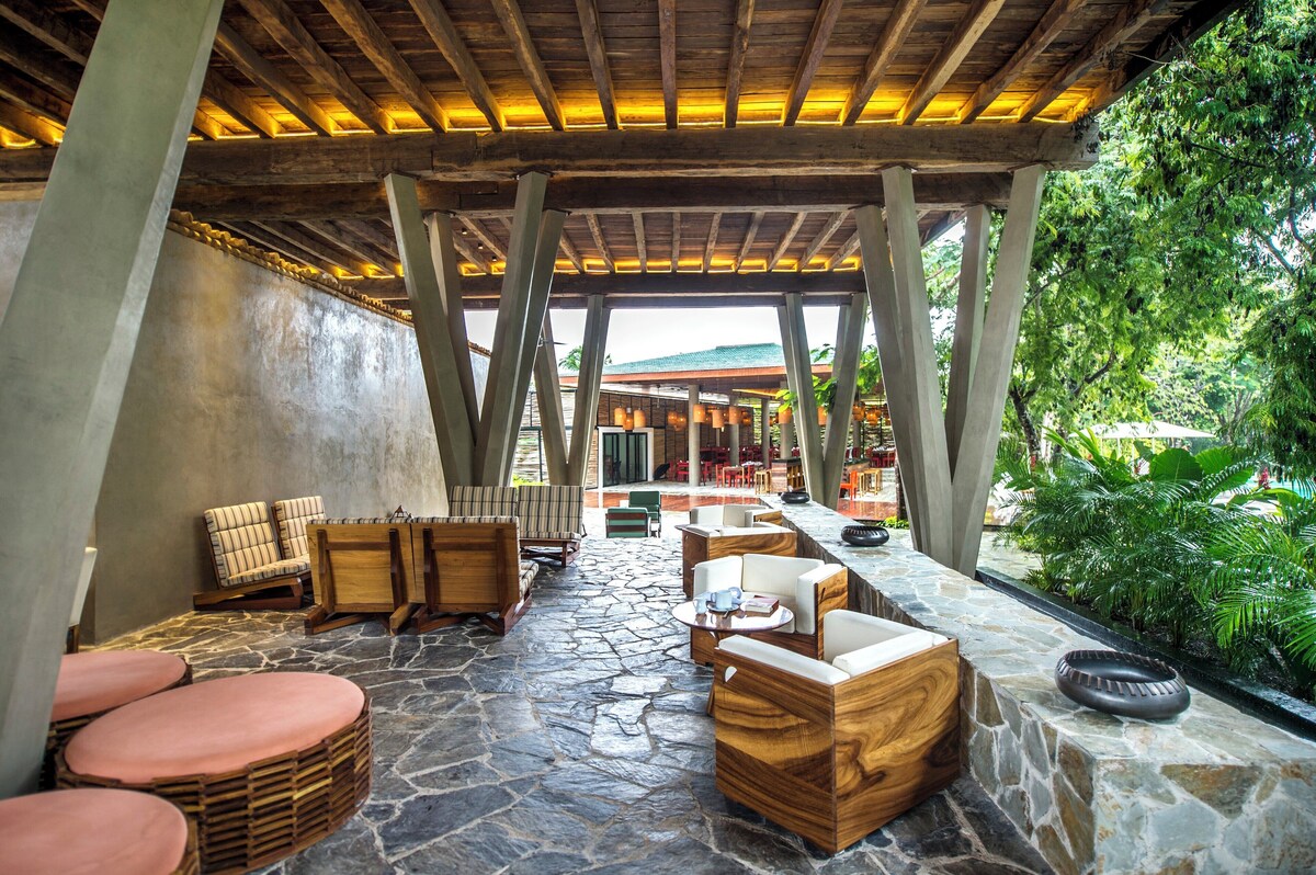 An outdoor seating area with stone flooring, wooden furniture, and a view of a building in the background. - The Explorean Cozumel - All Inclusive (Cozumel)
