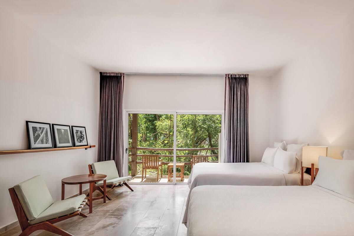 Une chambre d’hôtel avec deux lits, une petite table, deux chaises, un canapé et un balcon avec des rideaux.