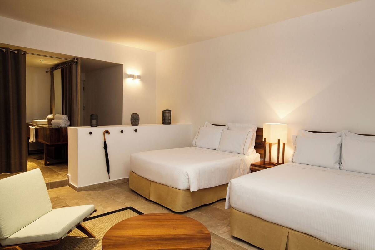 Une chambre d’hôtel avec deux lits, un coin salon comprenant une chaise et une petite table, une salle de bain avec un miroir et un porte-serviettes, et un luminaire fixé au mur.