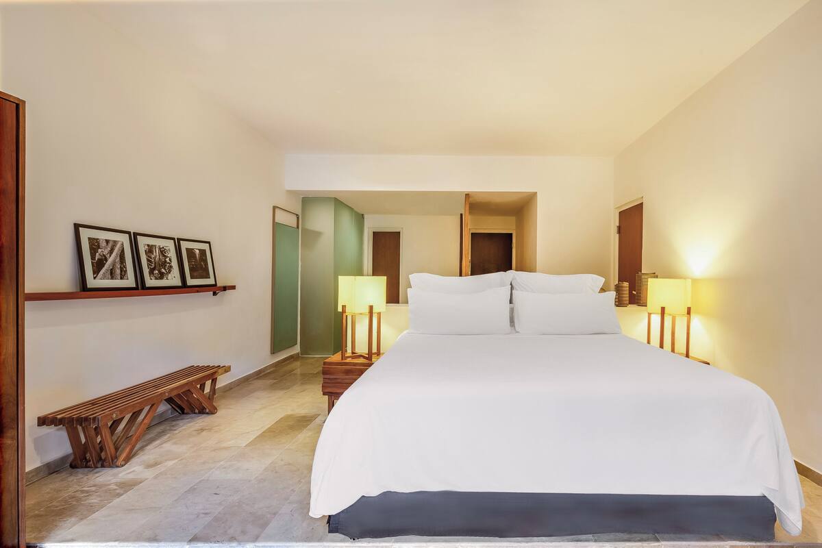 Une chambre d’hôtel avec un grand lit, deux lampes de chevet, un banc en bois et des tableaux encadrés au mur.