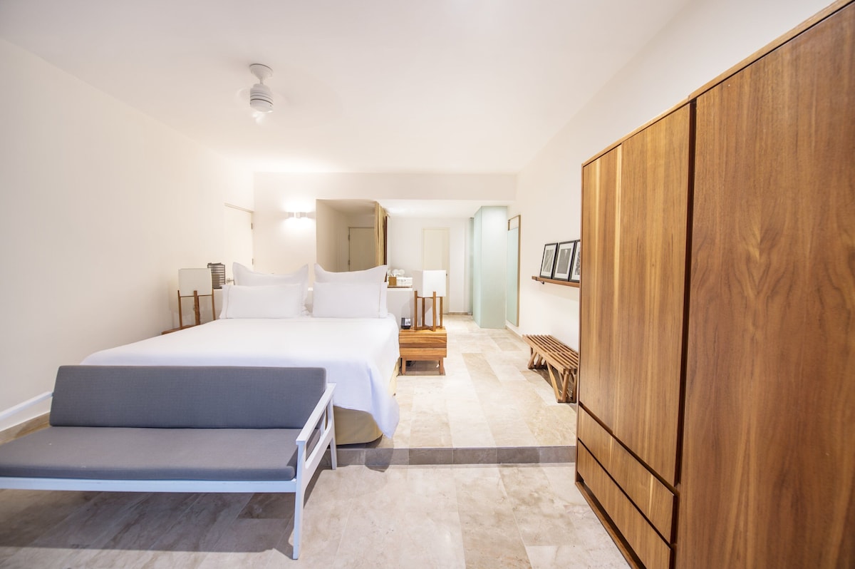 A modern hotel room with a large bed, a wooden wardrobe, a bench, and a nightstand with a lamp. - The Explorean Cozumel - All Inclusive (Cozumel)