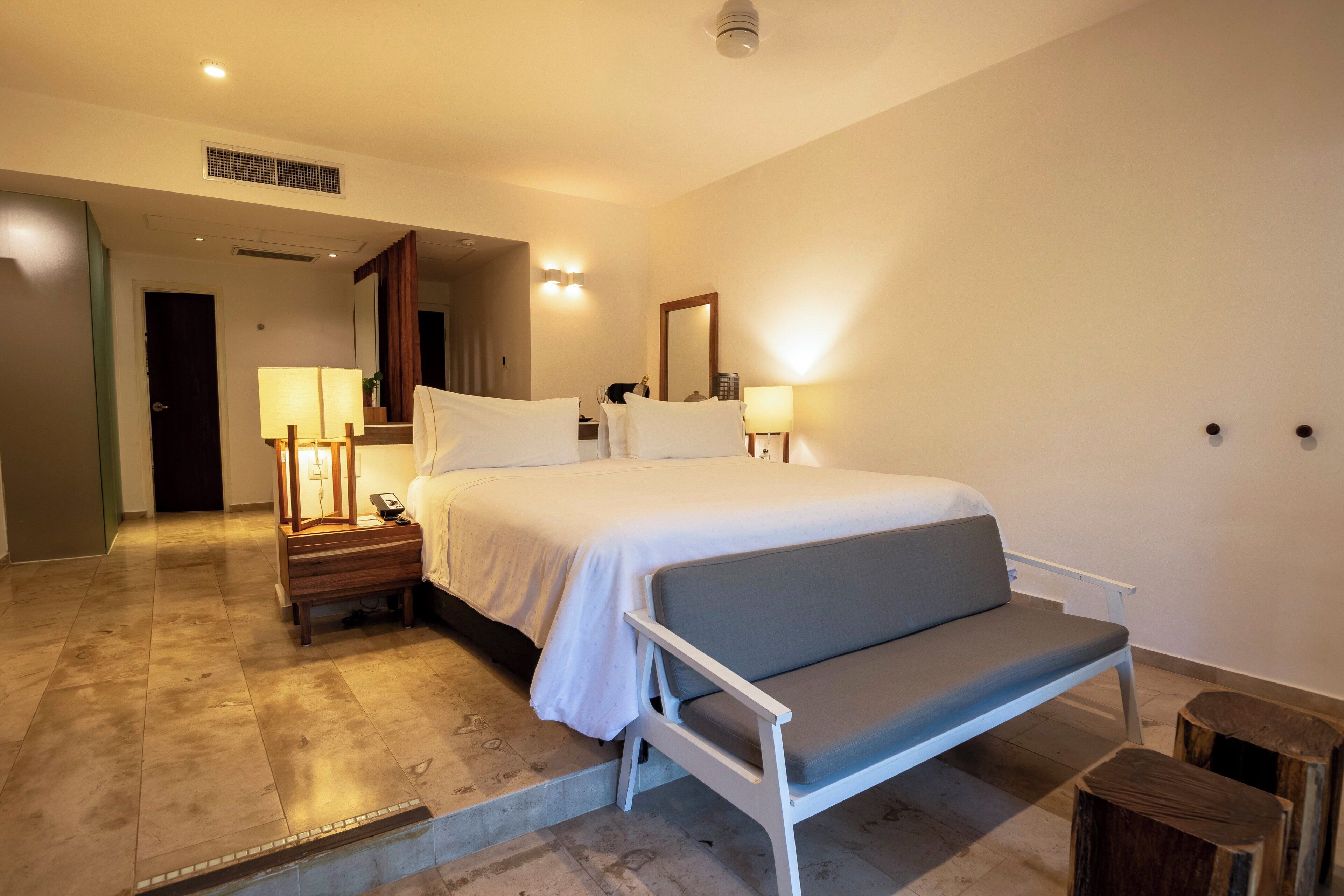 Una habitación de hotel con una cama, lámparas de noche, mesitas de noche, un banco y un taburete pequeño.
