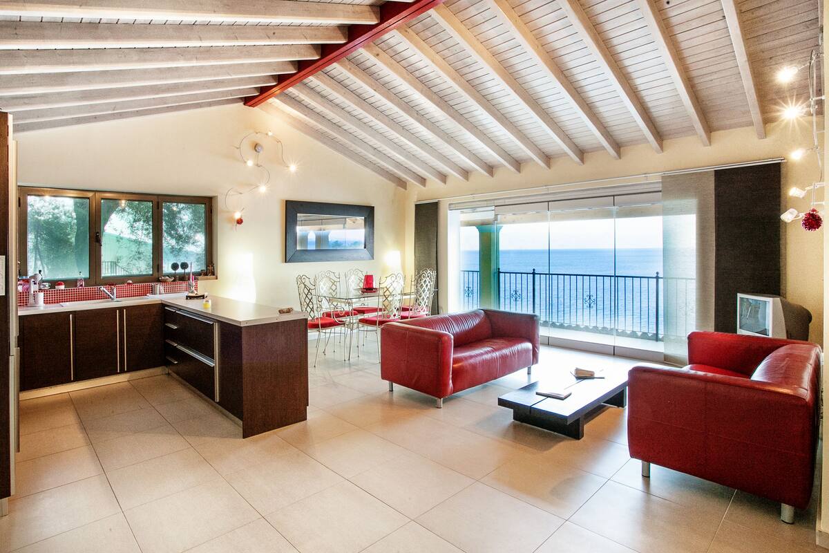 Amplia sala de estar con cocina, sofás de cuero rojo, una mesa de comedor y un gran ventanal con vistas al mar.