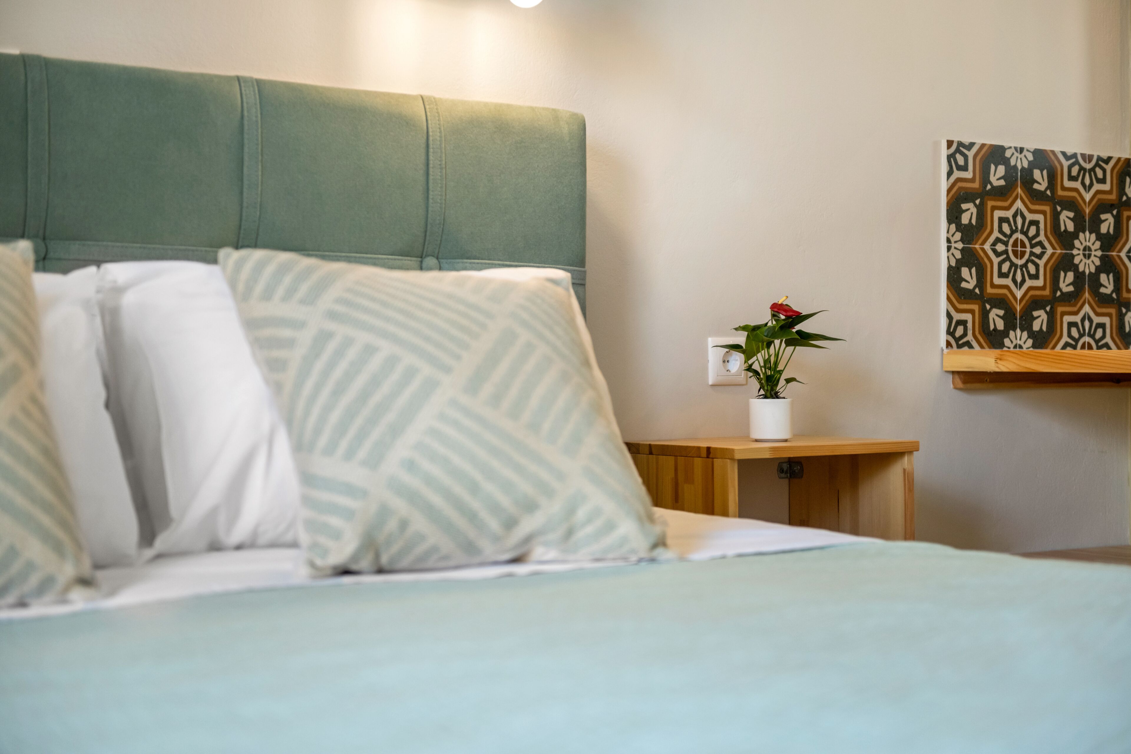 A neatly made bed with a green headboard, a bedside table with a potted plant, and a decorative tile on the wall.