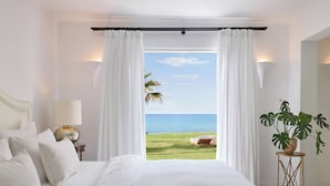 A bedroom with a large bed, white curtains, a view of the ocean, a potted plant, and a lamp on a bedside table.