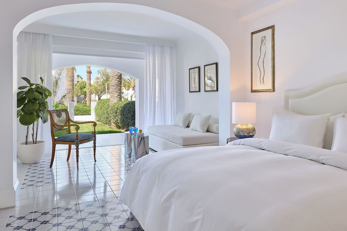 Habitación de hotel con cama, sofá, sillón y una mesita. Se ve un jardín a través de un arco.