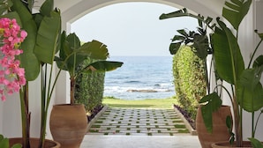 An arched pathway leading to a view of the ocean, flanked by large potted plants and a tiled walkway.