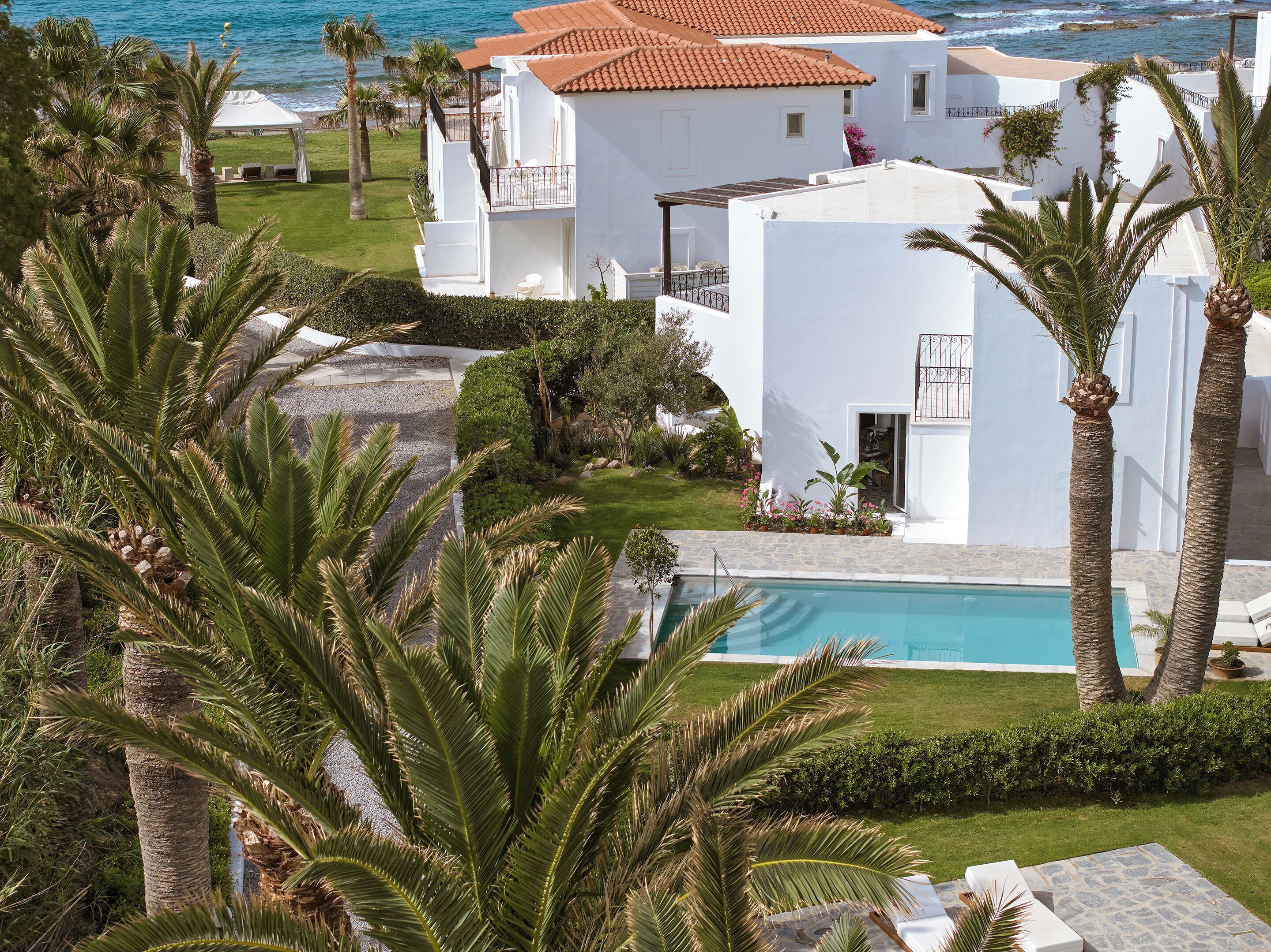 A white building with a pool, surrounded by palm trees and a view of the sea.