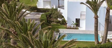A white building with a pool, surrounded by palm trees and a view of the sea.
