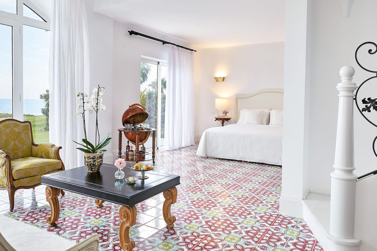 Habitación de hotel con cama, zona de estar con mesa, una silla y vista al mar.