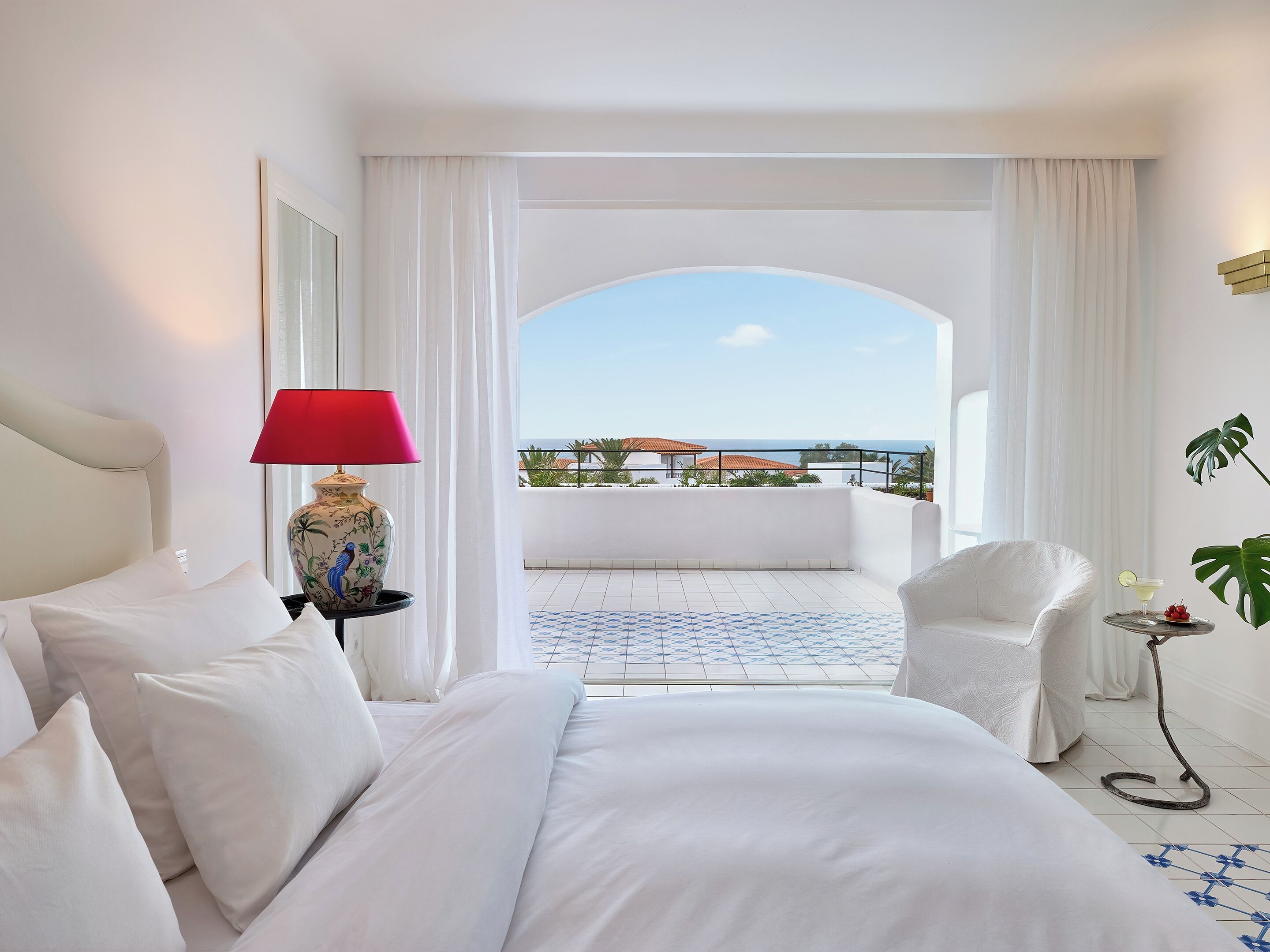 A hotel room with a bed, a red lamp, a chair, and a view of the sea.