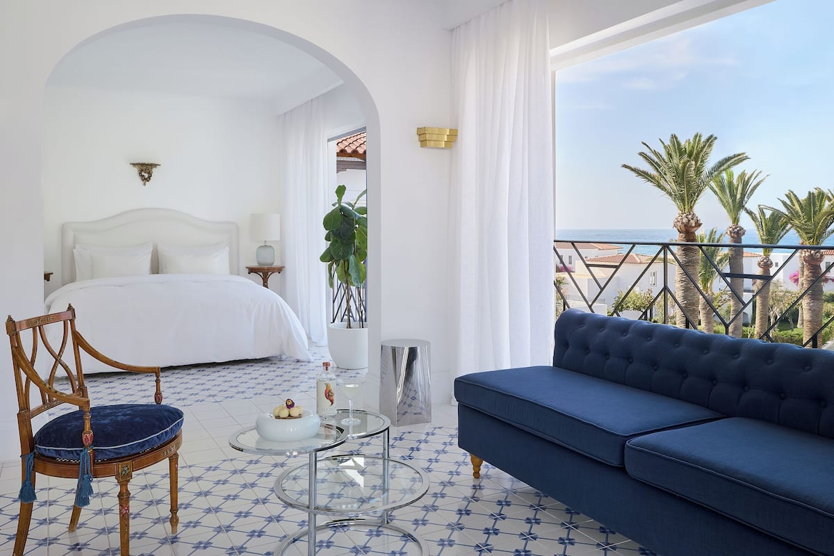 Habitación de hotel con cama, sofá azul, una silla, una mesita con una botella y un vaso, y vista al mar.