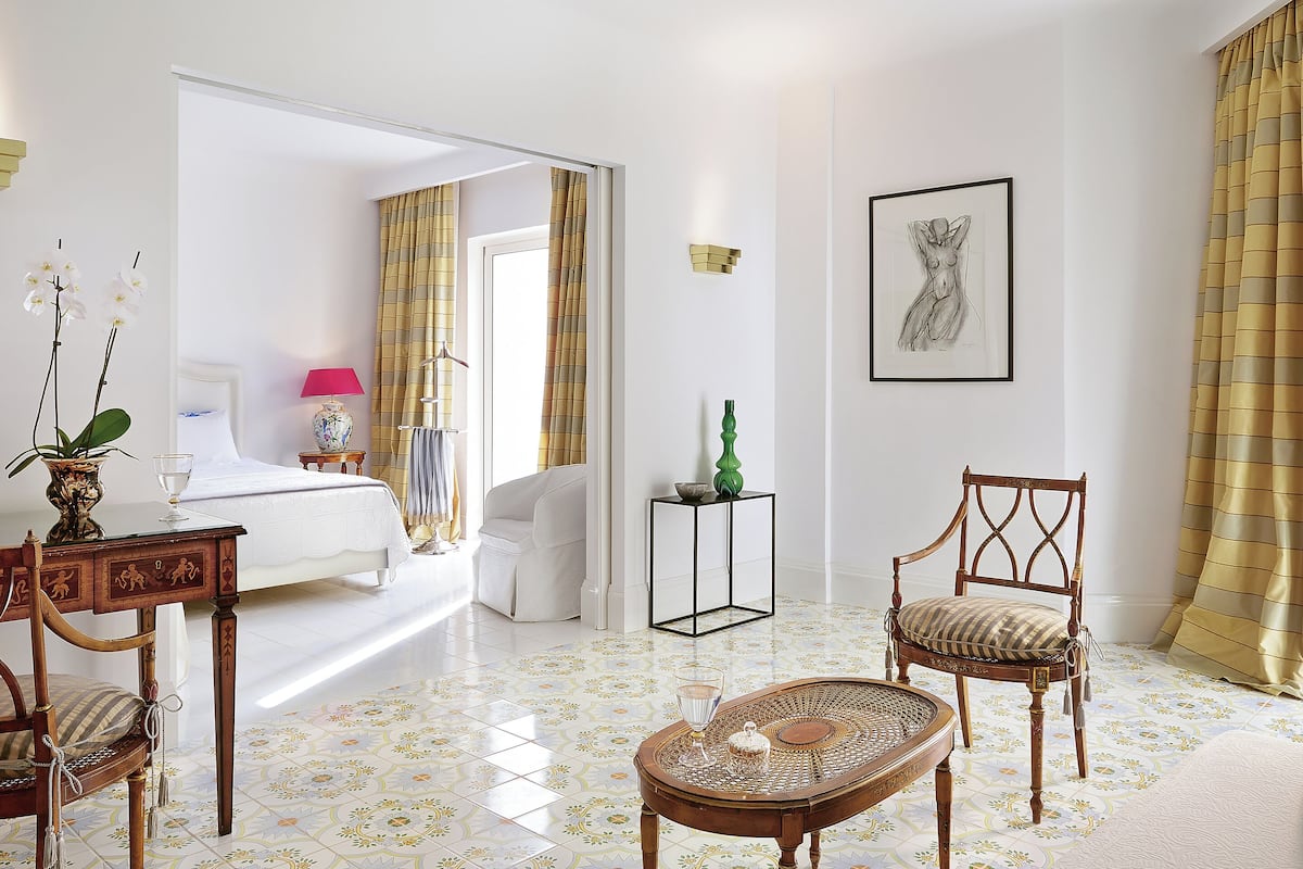 Una habitación de hotel con cama, una zona de estar con sillón y mesa, una mesita de noche con un florero, y una obra de arte enmarcada en la pared.