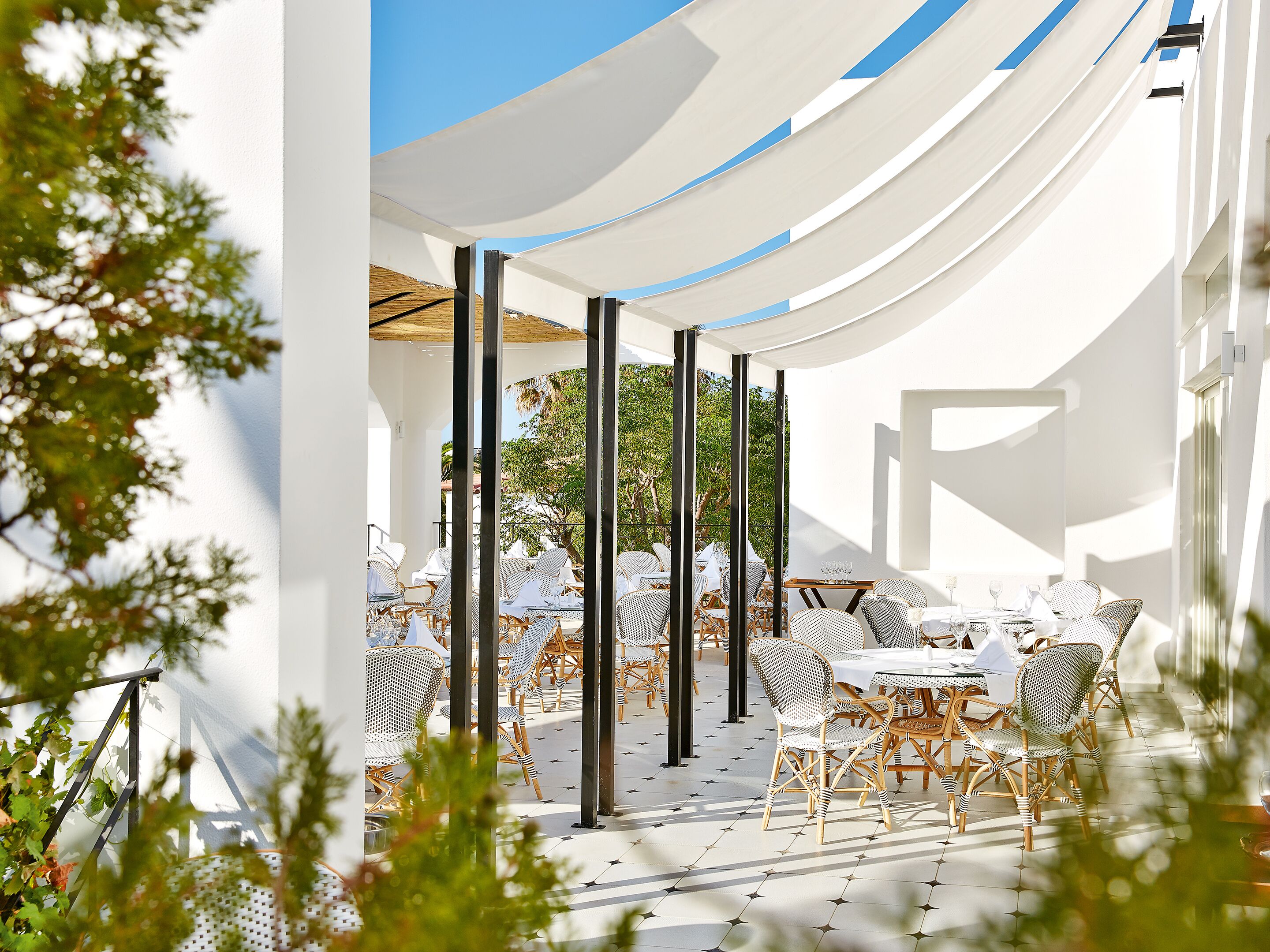An outdoor seating area with white tables and wicker chairs, shaded by white canopies, and surrounded by greenery.