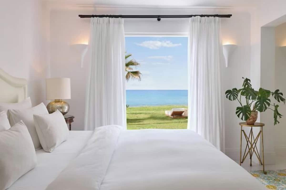 Un dormitorio con cama, cortinas blancas, una lámpara, una mesita, una planta en maceta y vistas al océano.