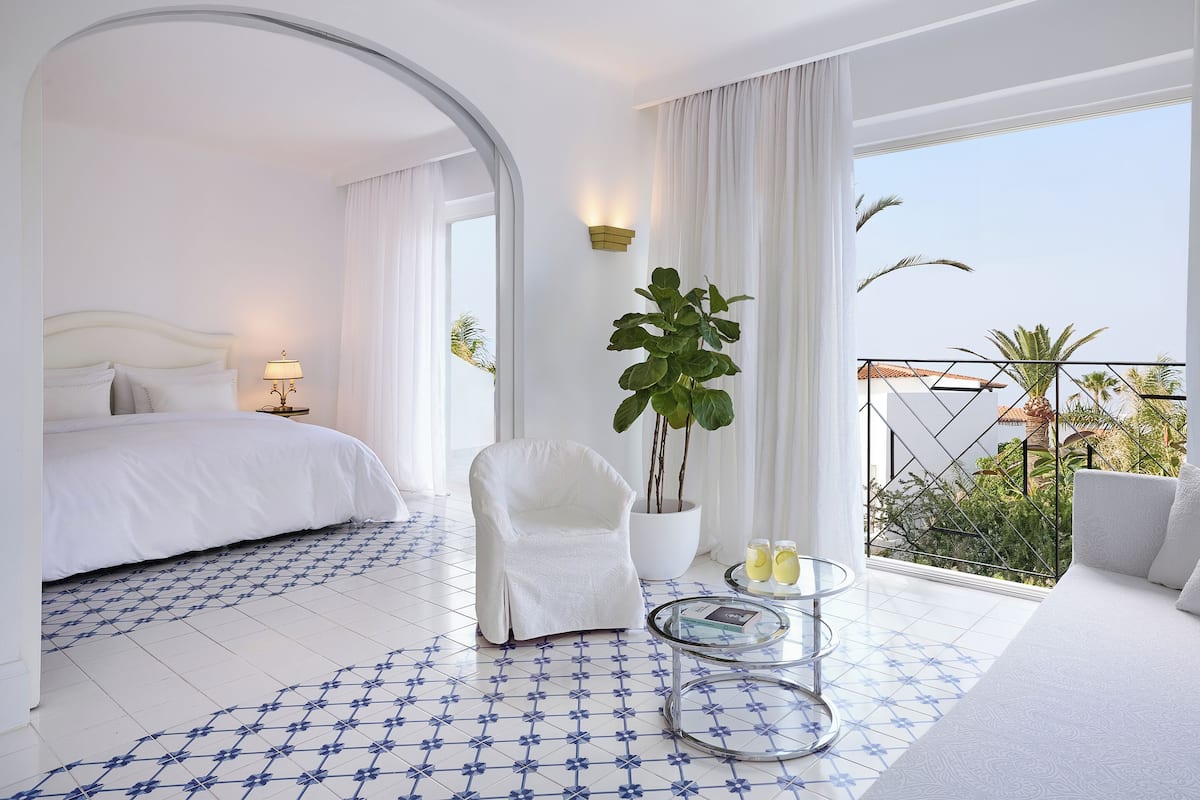 Habitación de hotel con cama, una silla, una mesita con bebidas, una planta en maceta y un balcón con vistas.