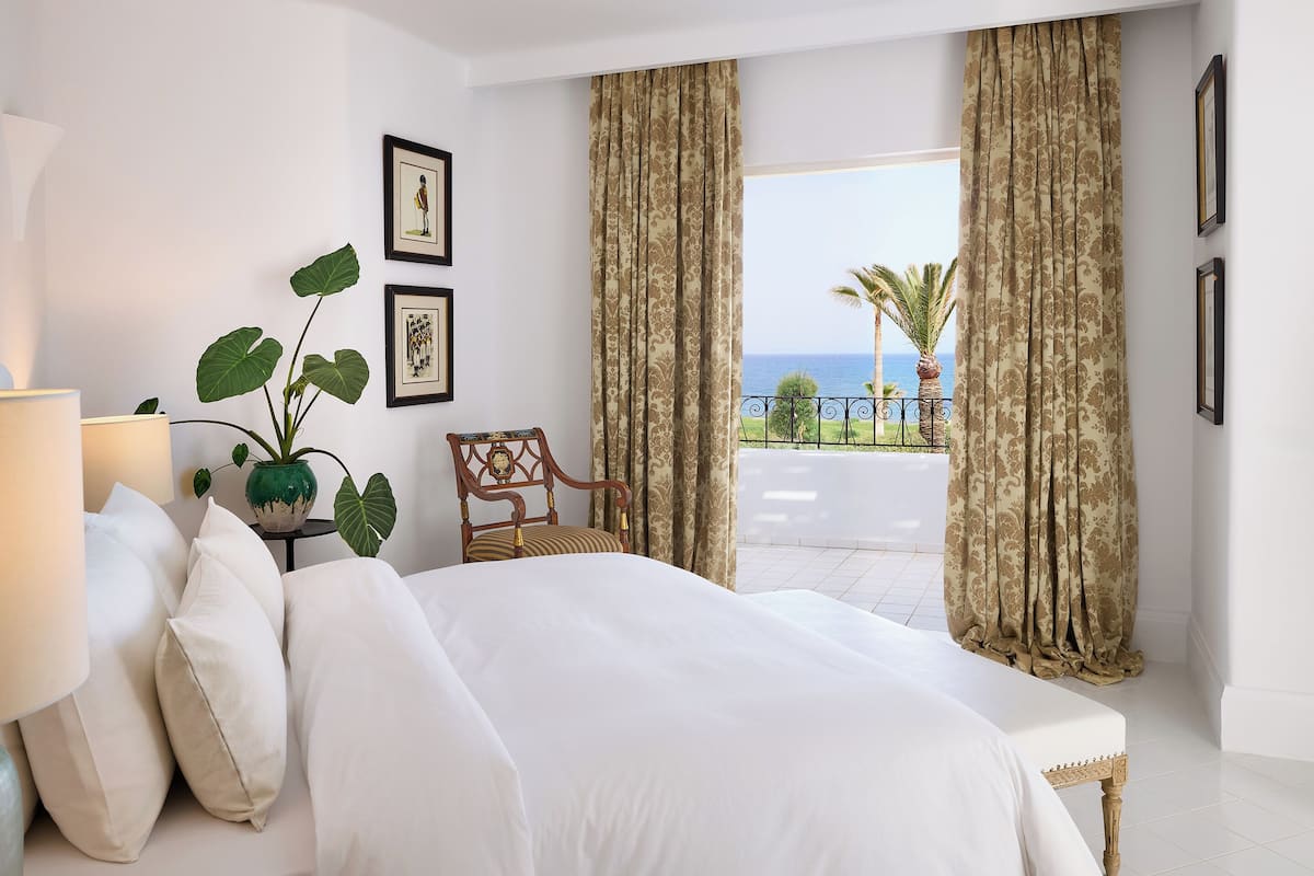 Un dormitorio con cama, una silla, una lámpara, una planta y cuadros en la pared.
