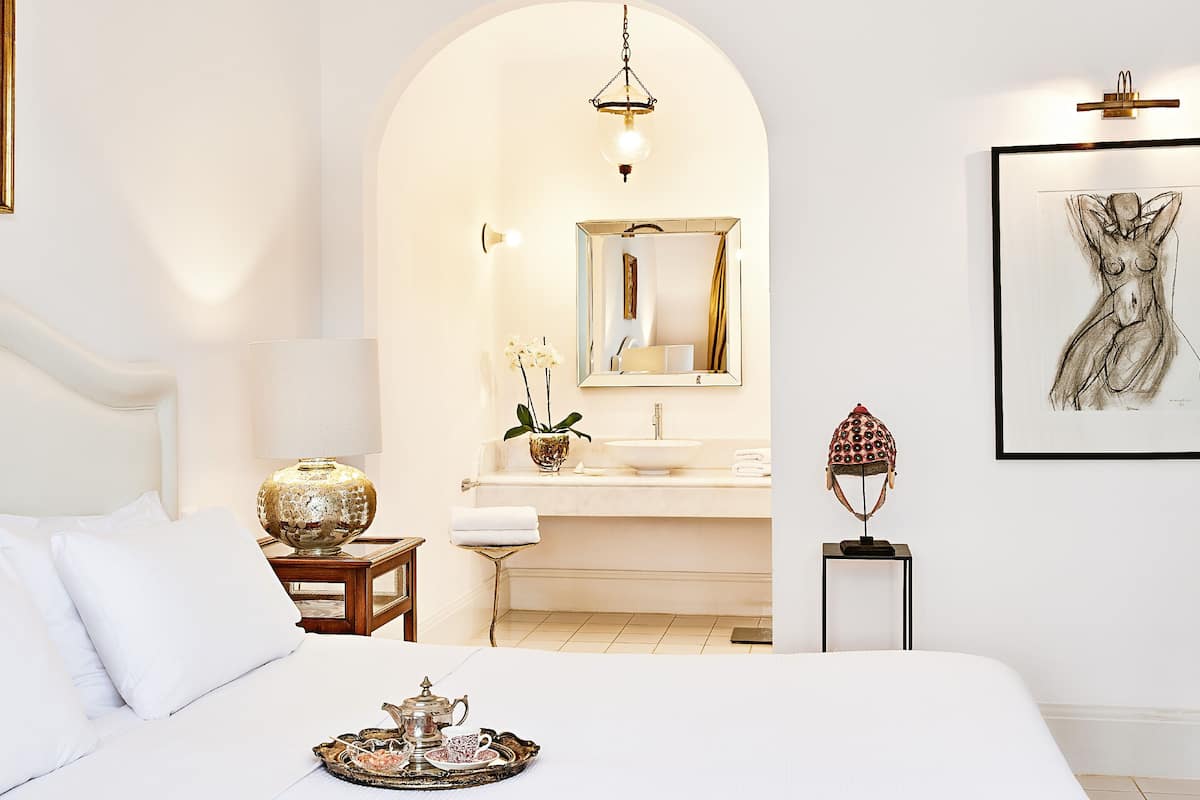 Un dormitorio con una cama grande, una mesita de noche con lámpara, una bandeja con juego de té, un baño con lavabo y espejo, y una obra de arte enmarcada en la pared.