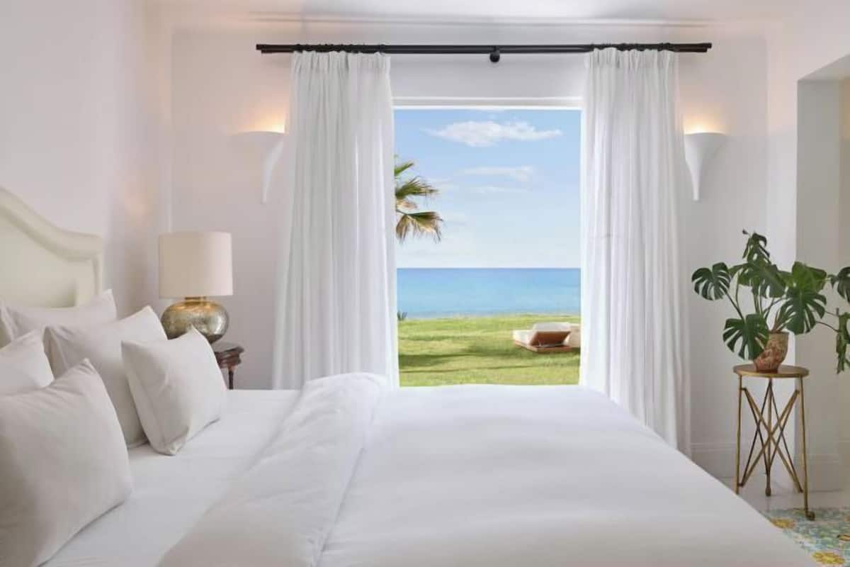 Un dormitorio con cama, cortinas blancas, vista al océano, una lámpara y una planta.