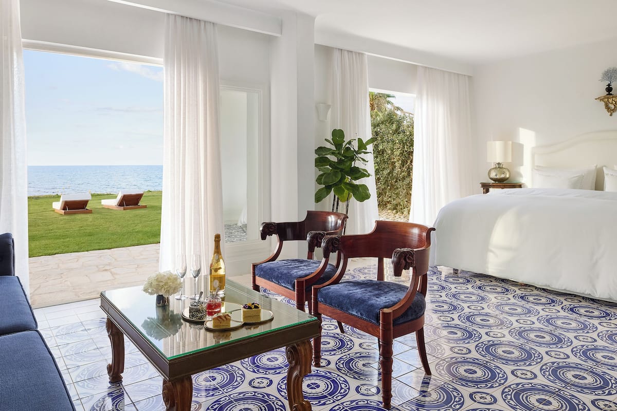 Habitación de hotel con una cama, una mesa con una botella y vasos, dos sillas, vista al mar y una planta grande.