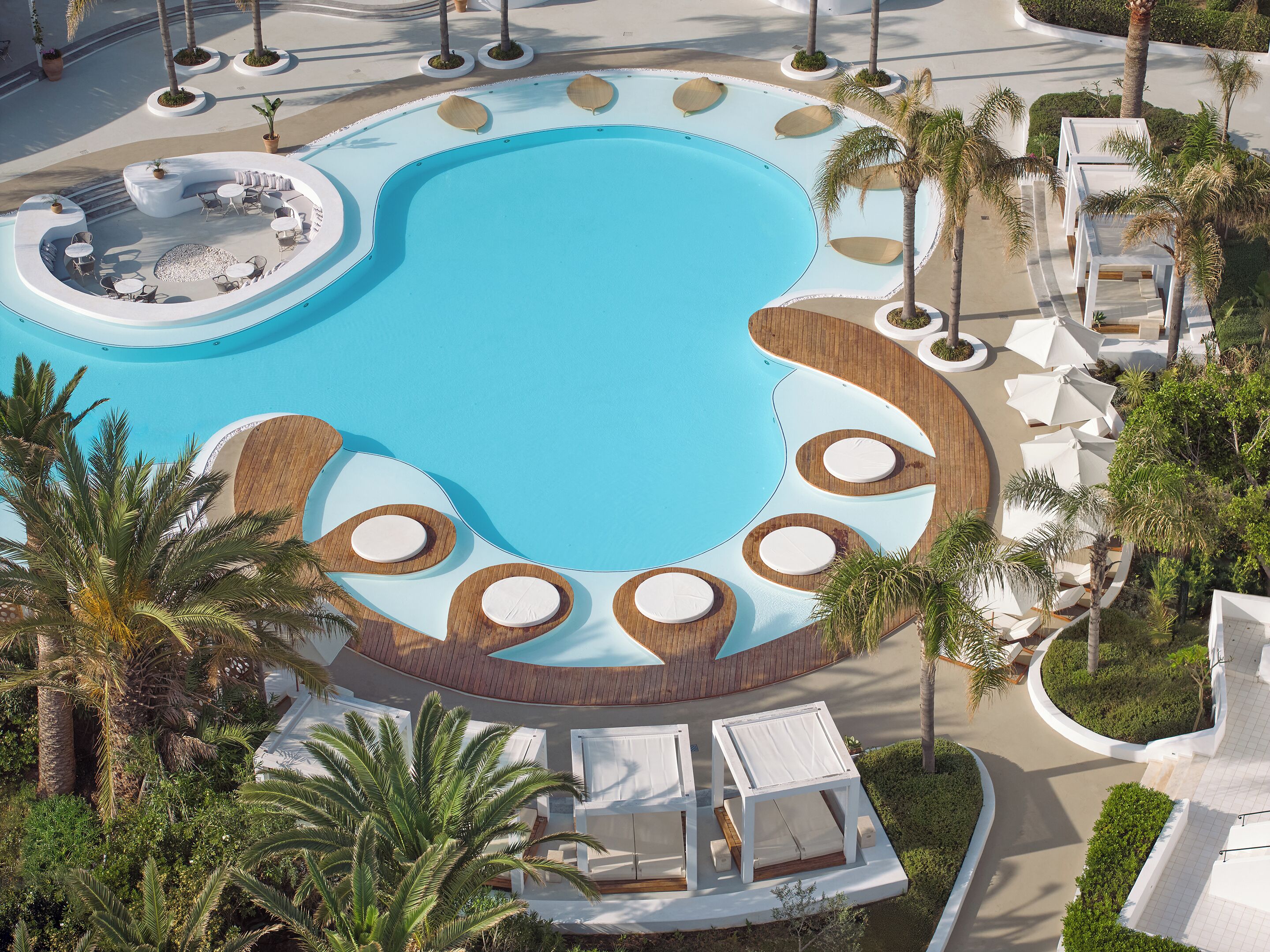 A large, circular swimming pool with multiple seating areas and palm trees.