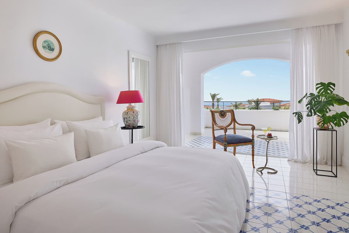 Habitación de hotel con una cama grande, una silla, una mesa con una bebida y vistas al mar.