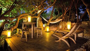 Una terraza de madera al aire libre con una mesa y sillas, rodeada de árboles e iluminada por faroles.