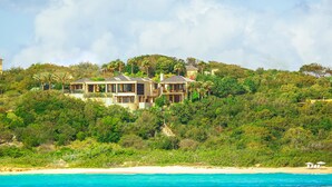Una casa grande de varios pisos con vista al mar, rodeada de abundante vegetación y árboles.