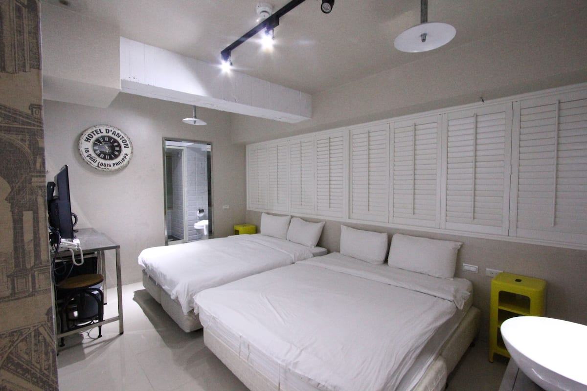 A hotel room with two beds, a desk with a computer, a small yellow stool, and a white sink.