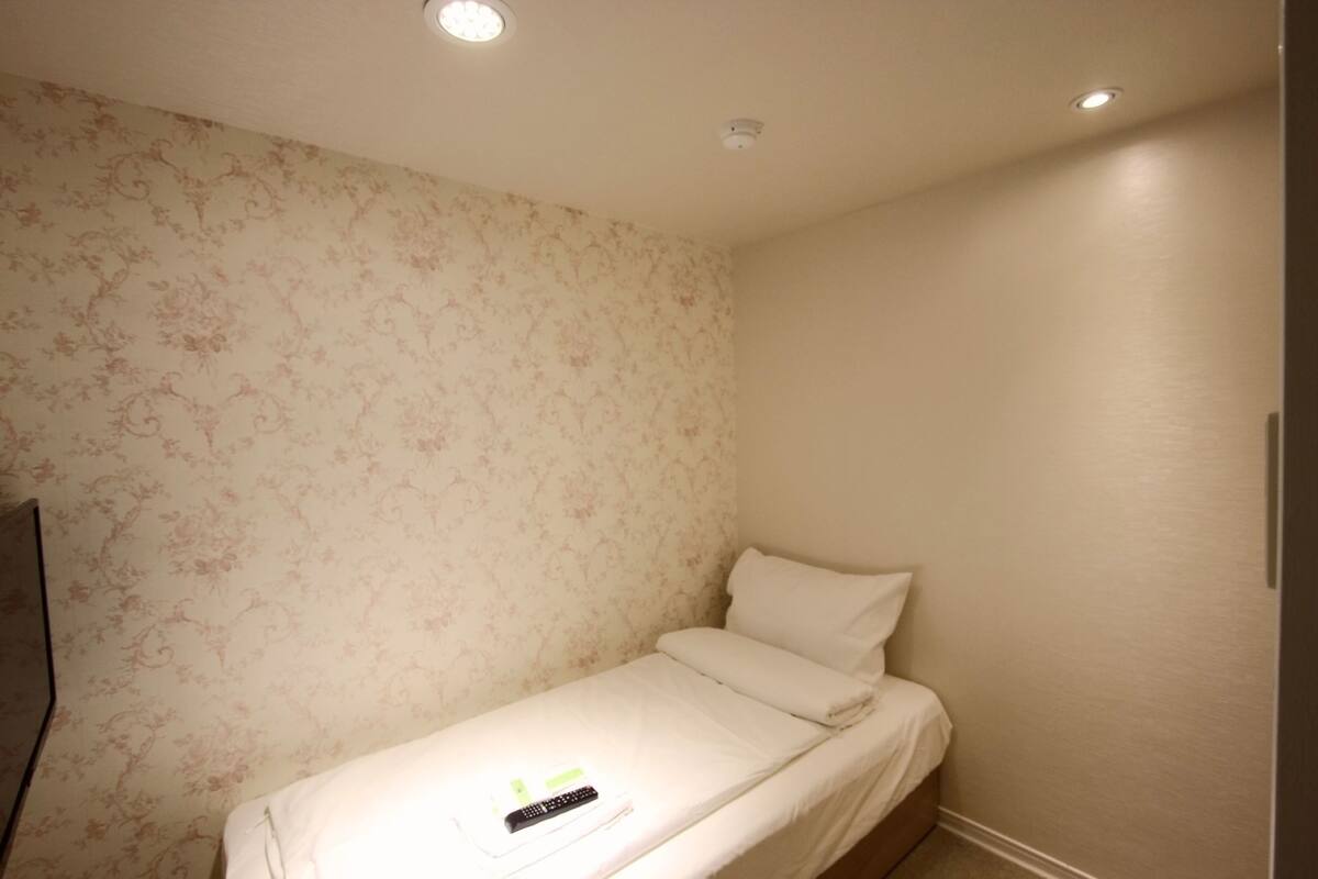 A small, single-bed room with floral wallpaper, a white bed, and a remote control on the bed.