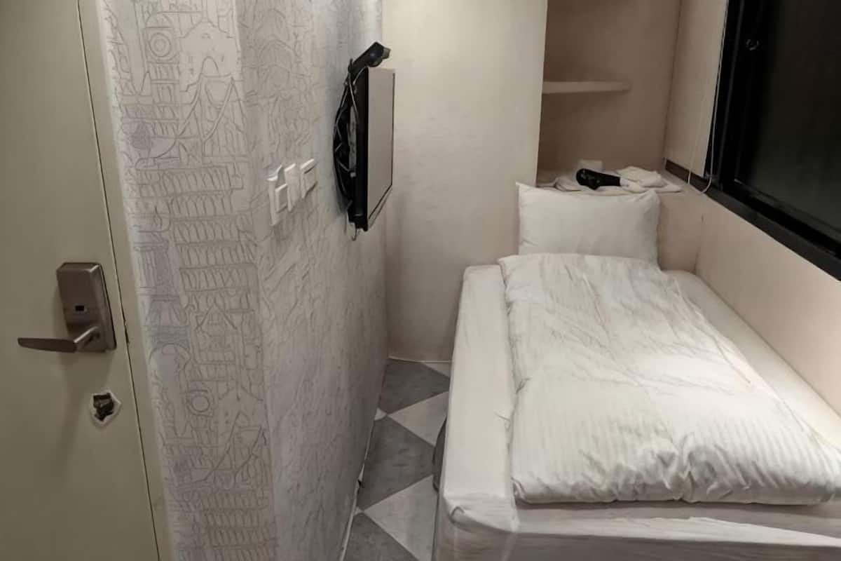A small, narrow room with a bed, a door with a handle, a wall-mounted light, and a window.