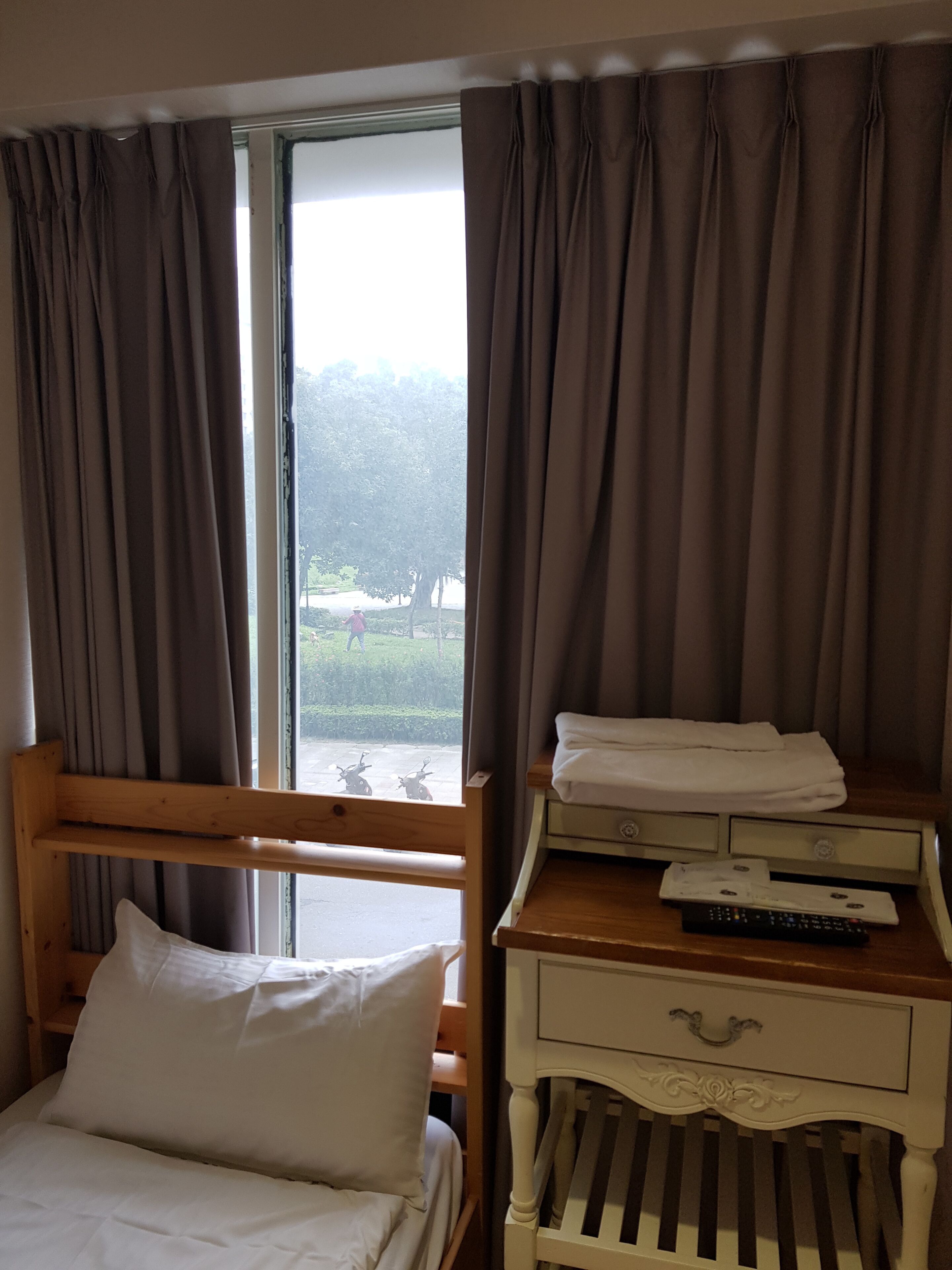 A hotel room with a bed, a wooden nightstand, a window with curtains, and a view of the outdoors.