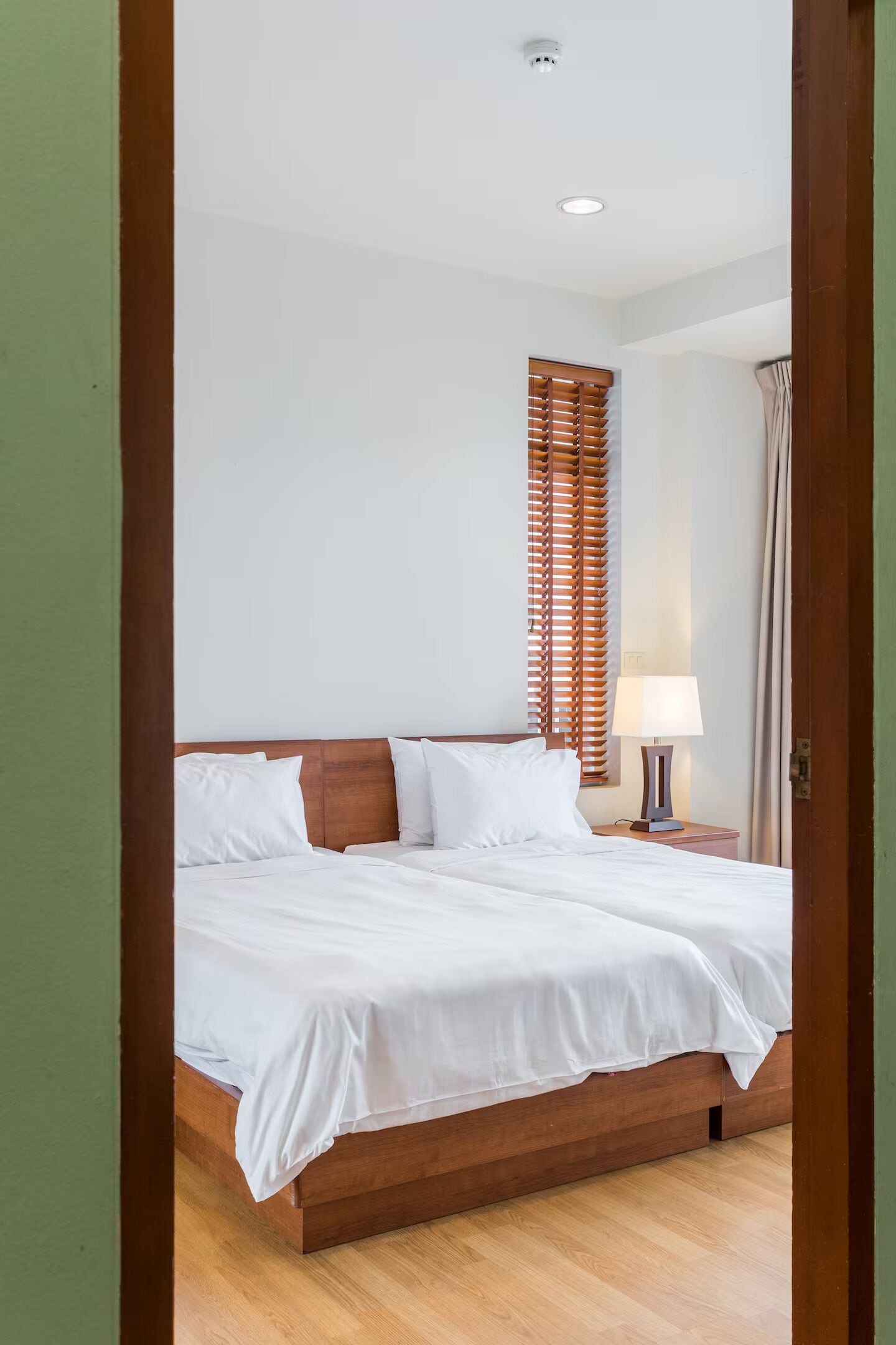 Un dormitorio con cama de madera, ropa de cama blanca, una mesita de noche con lámpara y una ventana con persianas.