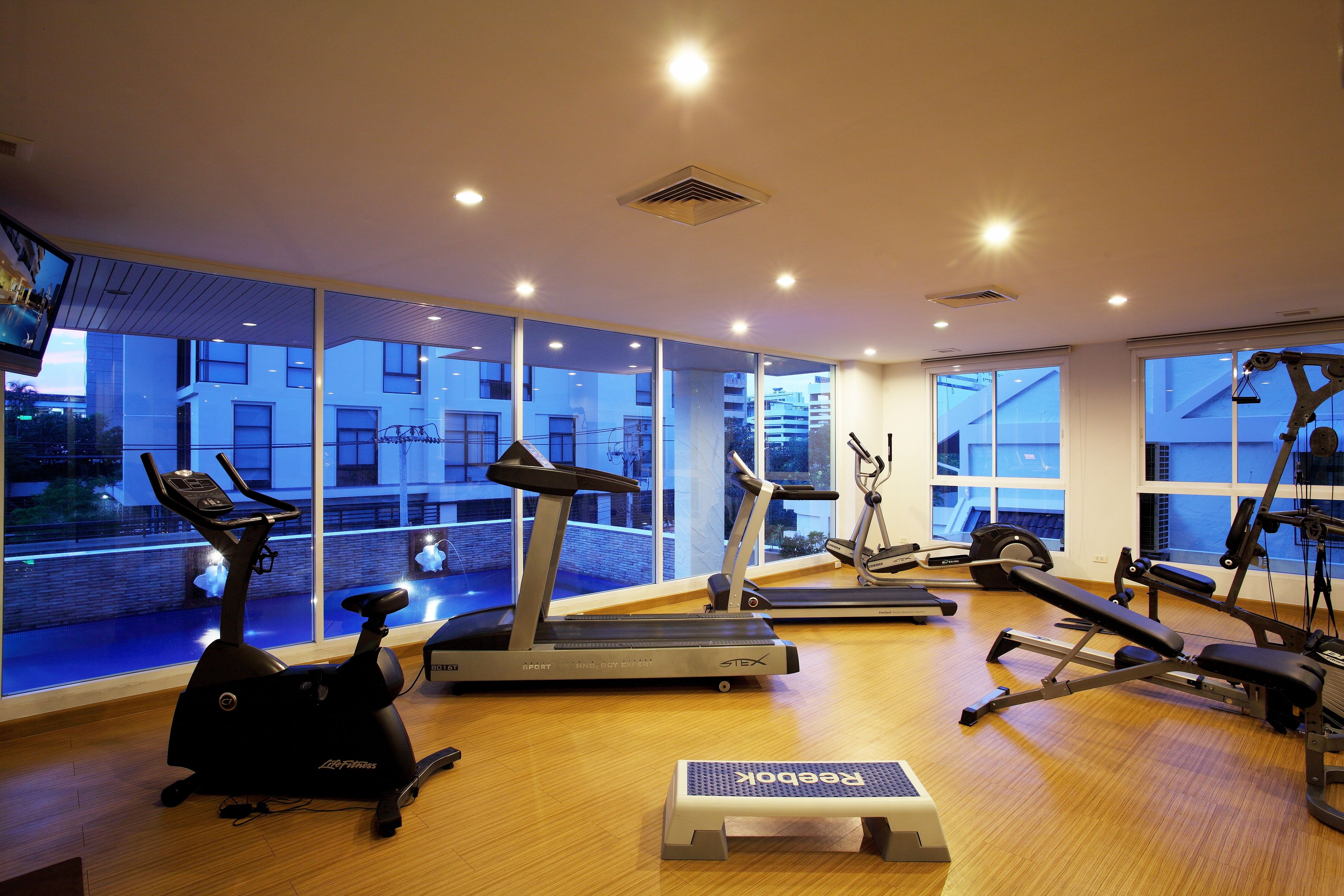 Ein Fitnessraum mit Laufbändern, Crosstrainern und einer Stepplatte, mit großen Fenstern, die einen Blick auf ein Gebäude und ein Schwimmbad bieten.