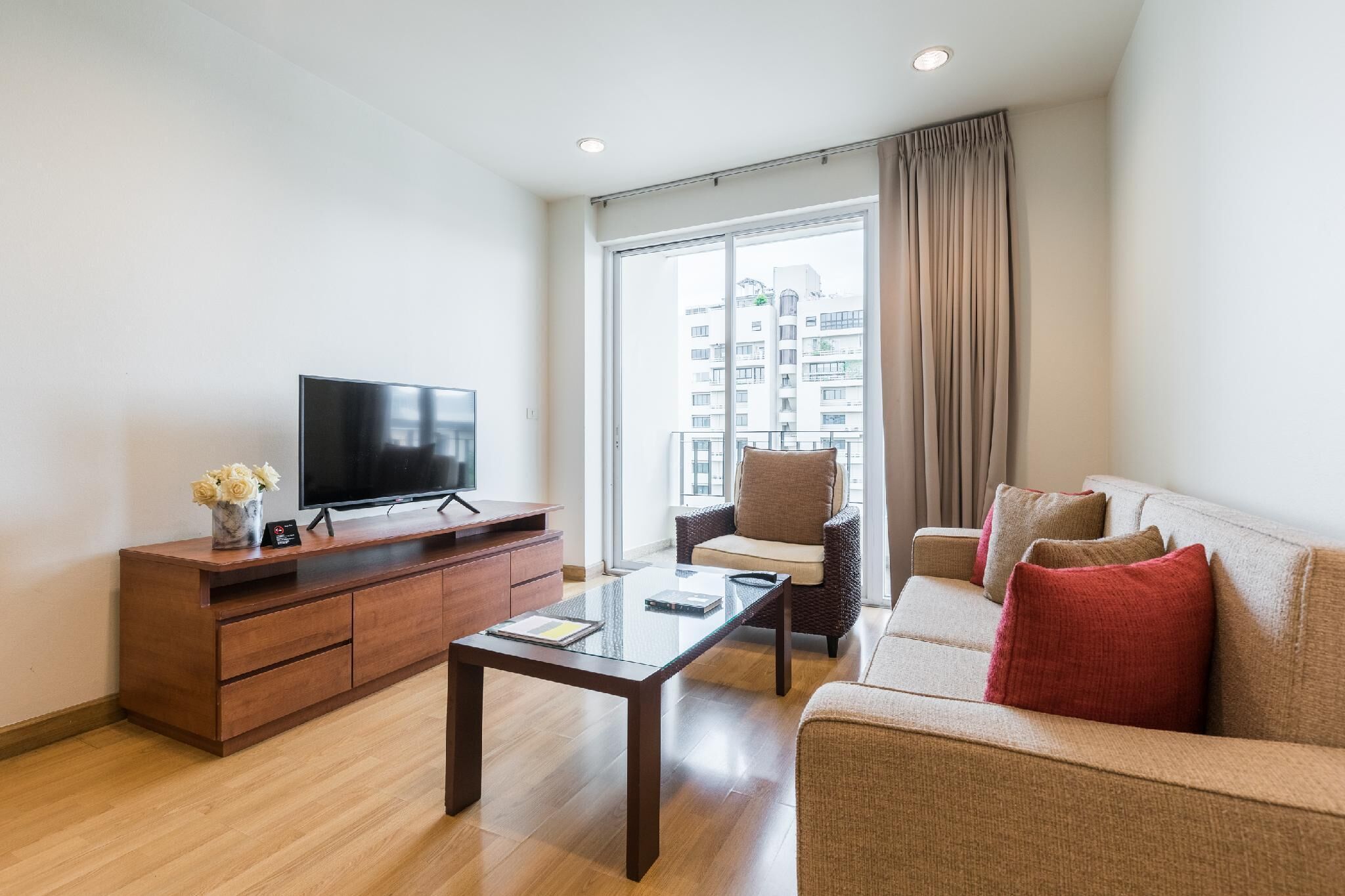 A modern living room with a flat-screen TV, wooden cabinet, glass coffee table, beige sofa, and a large window with curtains.