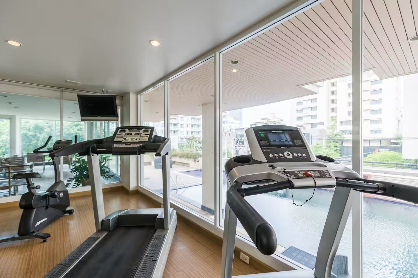 Un gimnasio con cintas de correr y una máquina elíptica, amplias ventanas y vistas a edificios y áreas verdes.