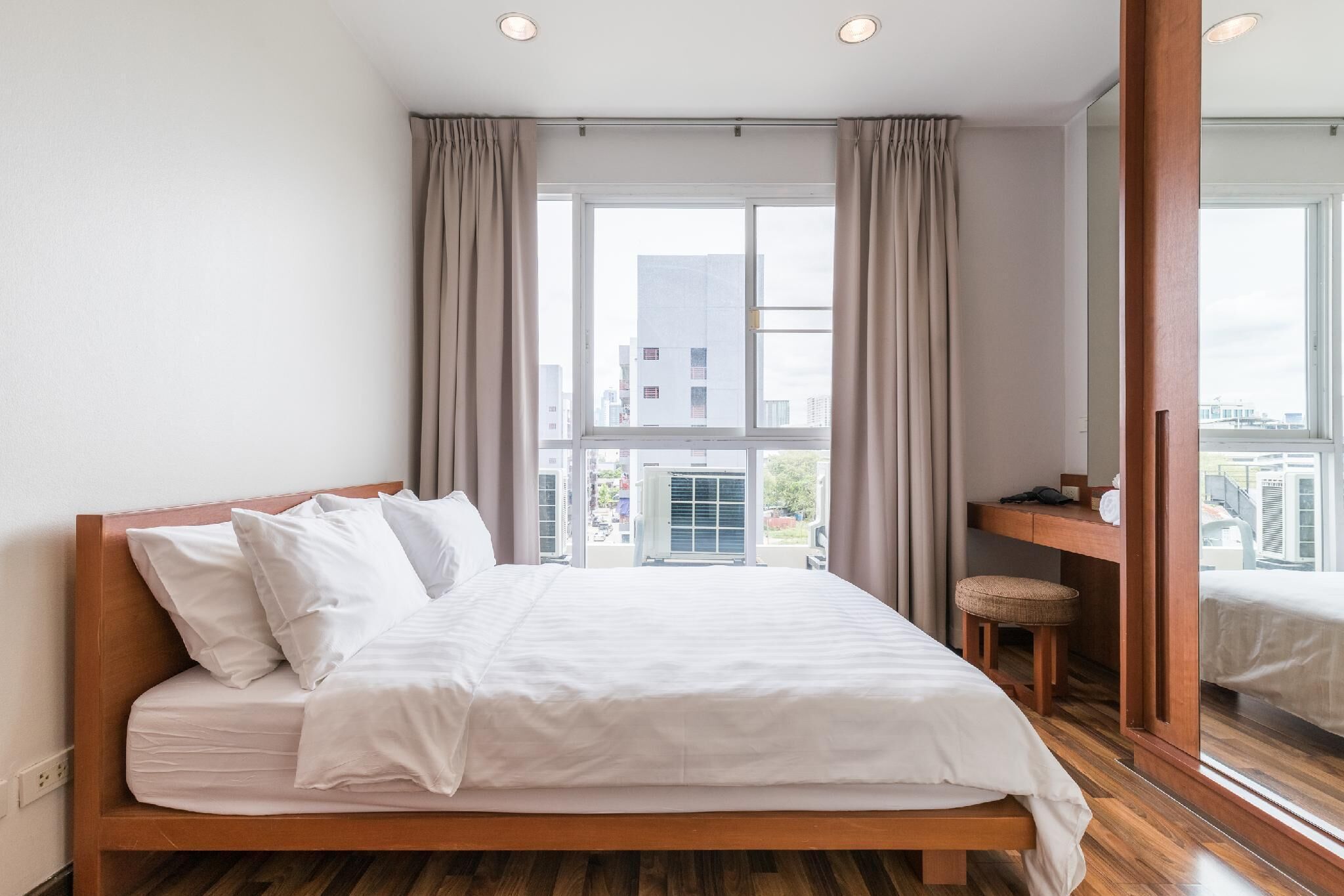 A modern hotel room with a wooden bed, a small desk, and a large window offering a city view.