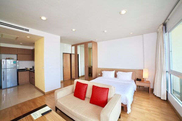 Une chambre d’hôtel avec un lit, un canapé avec des coussins rouges, une table de chevet avec une lampe, et une vue sur la cuisine et la salle de bain par une porte ouverte.